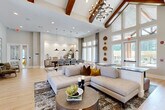 Located at 7001 Clementine Trl in Fairburn, GA, this single-floor community clubhouse offers a generous, open-concept layout designed for both comfort and functionality. The space is immediately defined by its dramatic vaulted cathedral ceilings accented with rich, exposed wood beams and a striking geometric chandelier, creating an impressive architectural focal point. Light-toned hardwood-style flooring runs throughout, complementing the bright, neutral color palette that keeps the interior feeling airy and welcoming. Expansive floor-to-ceiling windows line the walls, flooding the space with natural light and offering views of the surrounding outdoor areas and landscaping. The living area features a large, tufted sectional sofa anchored by a patterned area rug and a round wooden coffee table, providing an inviting lounge setting. Adjacent, the dining area is appointed with a solid wood table, woven counter-height chairs, and a trio of pendant lights overhead, alongside a sleek wet bar with tile backsplash detailing. A game and entertainment zone includes a pool table, wall-mounted televisions, and additional lounge seating. A hospitality station and a curated gallery hallway with framed artwork and accent seating round out the space, while decorative wood-slice wall art adds a distinctive organic touch throughout.