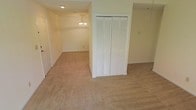 This single-floor space presents a clean, functional layout with neutral tones throughout. The flooring consists of wall-to-wall beige carpet, lending warmth and continuity across the open areas. A prominent built-in closet with classic white louvered bi-fold doors serves as a central storage feature, providing generous organizational capacity. The walls are finished in a soft, off-white palette that contributes to a bright and airy atmosphere. An adjoining room is visible in the background, distinguished by an elegant chandelier that adds a decorative focal point and overhead illumination to the space. Recessed ceiling lighting supplements the chandelier, ensuring consistent brightness throughout. The layout flows openly between areas, creating a sense of spaciousness. Door frames and trim are finished in white, complementing the overall neutral aesthetic. The space appears well-maintained and move-in ready, offering a versatile backdrop suitable for a variety of furnishing arrangements and uses. The single-floor design ensures easy accessibility and straightforward navigation throughout the home.