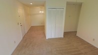 This single-floor space presents a clean, functional layout with neutral tones throughout. The flooring consists of wall-to-wall beige carpet, lending warmth and continuity across the open areas. A prominent built-in closet with classic white louvered bi-fold doors serves as a central storage feature, providing generous organizational capacity. The walls are finished in a soft, off-white palette that contributes to a bright and airy atmosphere. An adjoining room is visible in the background, distinguished by an elegant chandelier that adds a decorative focal point and overhead illumination to the space. Recessed ceiling lighting supplements the chandelier, ensuring consistent brightness throughout. The layout flows openly between areas, creating a sense of spaciousness. Door frames and trim are finished in white, complementing the overall neutral aesthetic. The space appears well-maintained and move-in ready, offering a versatile backdrop suitable for a variety of furnishing arrangements and uses. The single-floor design ensures easy accessibility and straightforward navigation throughout the home.