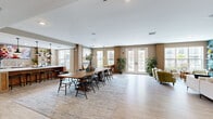 Located at 5885 Lakehurst Dr in Orlando, FL, this single-floor community amenity space offers a thoughtfully designed, open-plan layout ideal for socializing and gathering. The interior is finished with light wood-look tile flooring throughout, creating a cohesive and airy feel across the entire space.

The main gathering area features a large communal dining table with mixed seating, centered on a textured area rug and surrounded by oversized windows and French doors that invite abundant natural light and views of the outdoor surroundings. A dedicated lounge zone provides comfortable seating, including a tufted white sofa and olive-green accent chairs, arranged around a wall-mounted flat-screen TV set against a bold teal shiplap accent wall with open display shelving.

The kitchenette area is well-equipped with granite countertops, warm-toned cabinetry, open shelving, stainless steel appliances including a wall oven, a sink, and a textured tile backsplash — all anchored by a large kitchen island with bar seating. A cozy nook with a rustic wood bench, a round table, and pendant lighting adds a charming, intimate corner to the space. Shiplap wall paneling and carefully placed tropical greenery reinforce the relaxed, coastal-modern aesthetic throughout.