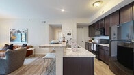 This thoughtfully designed 2-bedroom, 2-bathroom residence in Galloway, Ohio offers a comfortable and functional single-floor layout. The open-concept kitchen and living area serves as the heart of the home, featuring rich dark espresso cabinetry, granite-style countertops, a breakfast bar island with seating, black appliances, and wood-look flooring. A pantry provides additional storage, while the kitchen flows seamlessly into the living room, which is brightened by a glass patio door that opens to a private outdoor patio with exterior views. The carpeted living area offers a relaxed, inviting ambiance. The primary bedroom is generously sized, with soft carpet, a large window for natural light, and direct access to a walk-in closet. Its en-suite bathroom features a dark wood vanity with granite-style countertops, tile flooring, and a walk-in shower. The second bedroom is cozy and well-lit, served by a second full bathroom with matching finishes and a tub-shower combination. Additional highlights include five closets for ample storage, a dedicated laundry room, a utility room, and a hallway connecting all spaces efficiently. The neutral color palette throughout creates a clean, modern aesthetic.