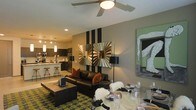 This single-floor space showcases an open-concept layout that seamlessly integrates the living, dining, and kitchen areas into one fluid environment. The living area is anchored by a sleek black leather sofa accented with rust-colored throw pillows, set atop a bold geometric-patterned area rug in olive green and charcoal tones. A ceiling fan with integrated lighting fixture sits overhead, complementing recessed lighting throughout. A dramatic accent wall featuring vertical black and silver stripes serves as a striking focal point, adorned with a sculptural starburst mirror that adds dimension and visual interest. Contemporary artwork on the adjacent wall further enhances the bold aesthetic. The dining area, positioned near the living space, features a glass-top table set with stylish place settings and white faux-fur chair accents, creating an eclectic yet curated atmosphere. The kitchen is visible in the background, outfitted with dark cabinetry, stainless steel appliances, a peninsula with bar-height seating, and pendant lighting hanging above. The overall ambiance is modern and design-forward, with a cohesive color palette of black, gray, olive, and warm orange tones.