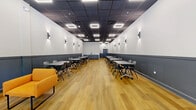 Located at 1 Richmond St in New Brunswick, NJ, this well-appointed single-floor space offers a thoughtfully designed interior suited for a range of uses, from meetings and workshops to social gatherings. The open layout is anchored by warm-toned hardwood flooring that runs the full length of the room, creating an inviting and cohesive aesthetic throughout.

The walls feature a two-tone design — crisp white upper panels paired with a deep charcoal wainscoting — lending a polished, contemporary feel. A dark drop ceiling with recessed square LED light fixtures provides even, modern illumination, while angular wall-mounted sconces add an additional layer of ambient lighting along the perimeter.

Multiple rectangular tables with mobile chairs are arranged in rows down the center and along the walls, offering flexible seating configurations to accommodate a variety of event formats. A comfortable lounge area near the entrance features bold orange upholstered sofas and chairs centered around a sleek chrome accent table, providing a welcoming and casual gathering spot.

Natural light filters in through glass-panel entry doors at one end of the room, and a utility counter with storage cabinetry is positioned along the rear wall. The overall ambiance is clean, functional, and modern, making this space well-suited for professional and community use alike.