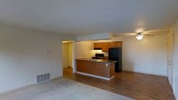 This single-floor residence offers an open-concept design that connects the living and kitchen areas in a functional and cohesive layout. The living area features neutral carpet flooring, a ceiling fan for year-round comfort, and a floor vent for efficient climate control, creating a relaxed and practical atmosphere. The kitchen is positioned at the heart of the space, anchored by a raised peninsula with a dark stone countertop that serves as both a workspace and a casual dining or gathering spot. Warm wood-toned cabinetry lines the upper walls, providing ample storage, while black appliances — including a refrigerator and range — add a bold contrast. The kitchen transitions smoothly into a hardwood-style floor, distinguishing it from the carpeted living zone. A hallway visible beyond the kitchen suggests access to additional rooms, further extending the functionality of the home. The white walls and standard-height ceilings keep the space feeling bright and open throughout. Overall, the layout prioritizes ease of movement and everyday practicality, making it a well-organized and accessible living environment.