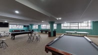 Located at 34 Franklin St in Nashua, NH, this single-floor amenity space offers a well-rounded recreational environment designed for community gathering and leisure. The expansive game room is a centerpiece of the floor, featuring multiple full-size billiard tables, a foosball table, and patterned carpeting underfoot. Distinctive teal-painted structural columns punctuate the open layout, adding a bold design accent throughout. Large industrial-style windows line the walls, flooding the space with natural light and offering views of the surrounding cityscape.

A comfortable lounge area provides rich burgundy leather sofas arranged around a coffee table, with glass partition walls offering sightlines into adjacent spaces, including a fitness area equipped with exercise machines. The dining and seating zones feature dark wood counter-height chairs positioned along windowed walls, creating inviting spots for socializing.

The kitchenette is outfitted with white cabinetry, granite countertops, stainless steel appliances including a refrigerator and dishwasher, and hardwood-style flooring — providing practical amenities for events or everyday use. An open mezzanine overlook with black metal railings connects the spaces visually, reinforcing the building's industrial-chic character throughout.