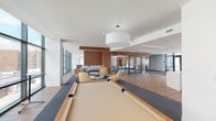 The Club Room at 360 Huguenot Street in New Rochelle, NY is a thoughtfully designed communal amenity space that blends modern aesthetics with comfortable, functional living. The open-plan layout is anchored by warm wood-paneled accent walls that provide a rich contrast against crisp white ceilings and light-toned flooring throughout.

At the heart of the lounge area, plush tan leather lounge chairs surround a sculptural round coffee table, creating an inviting social hub. A striking double-sided glass fireplace, framed in warm wood paneling, serves as an elegant divider between the seating zones. Floor-to-ceiling windows flood the space with natural light and offer expansive outdoor views, enhancing the bright, airy atmosphere.

A full-size billiards table anchors one end of the room beneath a large pendant drum light, adding a recreational focal point. Adjacent to the main lounge, a glass-enclosed dining and meeting area features multiple seating arrangements and direct access to an outdoor terrace through full-height glass doors.

The kitchen and bar area is outfitted with a generous white countertop island with modern bar stools, paired with warm wood cabinetry and a full run of lower cabinetry with integrated appliances — ideal for entertaining. The overall ambiance is polished, modern, and welcoming, making this Club Room a premier gathering space for residents.