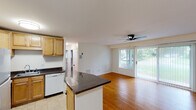 This single-floor unit offers a functional and well-connected layout that seamlessly blends the kitchen and living areas into one open space. The kitchen is equipped with warm-toned maple cabinetry, dark granite countertops, a white dishwasher, a refrigerator, and a sink, all set over light tile flooring. A peninsula counter extends into the living area, creating a natural divide while maintaining an open feel. The living room is bright and spacious, featuring warm hardwood flooring, a ceiling fan, and large sliding glass doors dressed with vertical blinds that flood the space with natural light and provide views of the lush greenery outside. One accent wall in the living area is clad in white square tile, adding a distinctive architectural detail. The two bedrooms continue the hardwood parquet flooring theme and are filled with natural light through large windows with horizontal blinds, offering views of the surrounding landscape. The bathroom is finished with clean white square tile surrounding a full bathtub and shower combination, complemented by a pedestal sink. Throughout the unit, neutral white walls and consistent flooring choices create a cohesive and bright atmosphere that is both practical and inviting.
