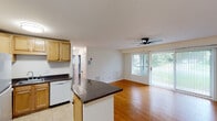 This single-floor unit offers a functional and well-connected layout that seamlessly blends the kitchen and living areas into one open space. The kitchen is equipped with warm-toned maple cabinetry, dark granite countertops, a white dishwasher, a refrigerator, and a sink, all set over light tile flooring. A peninsula counter extends into the living area, creating a natural divide while maintaining an open feel. The living room is bright and spacious, featuring warm hardwood flooring, a ceiling fan, and large sliding glass doors dressed with vertical blinds that flood the space with natural light and provide views of the lush greenery outside. One accent wall in the living area is clad in white square tile, adding a distinctive architectural detail. The two bedrooms continue the hardwood parquet flooring theme and are filled with natural light through large windows with horizontal blinds, offering views of the surrounding landscape. The bathroom is finished with clean white square tile surrounding a full bathtub and shower combination, complemented by a pedestal sink. Throughout the unit, neutral white walls and consistent flooring choices create a cohesive and bright atmosphere that is both practical and inviting.