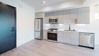 This single-floor space showcases a clean, contemporary design throughout. The open-concept layout is unified by light wood-toned flooring that extends seamlessly across the living areas and bedrooms, creating a bright and cohesive aesthetic.

The kitchen is well-appointed with floor-to-ceiling light wood cabinetry, stainless steel appliances including a gas range, microwave, refrigerator, and dishwasher, complemented by a geometric white tile backsplash and light stone countertops. Recessed lighting and under-cabinet fixtures ensure the space is well-lit.

The living area benefits from large sliding glass doors that open to a private balcony, offering views of surrounding greenery and filling the room with natural light. Recessed ceiling lights add to the bright, airy ambiance.

The bedrooms feature crisp white walls, recessed lighting, and ceiling fans. One bedroom includes a built-in closet with frosted sliding glass doors, adding a refined touch. Additional rooms offer ample wall space and multiple entry points.

The bathroom presents a polished finish with large-format marble-style porcelain floor tiles, a floating wood-toned vanity, an illuminated mirror, and a clean white shower area, all tied together with modern chrome fixtures.