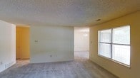 This single-story space presents a straightforward, functional layout with a clean and neutral aesthetic throughout. The main living area is generously sized, featuring wall-to-wall carpet flooring in a light gray tone that contributes to a calm, understated atmosphere. The walls are finished in a soft beige, providing a versatile backdrop suitable for a variety of interior styles.

The ceiling features a classic popcorn texture, a common characteristic of its construction era, with a centrally located HVAC vent ensuring climate comfort. A window fitted with horizontal blinds on the far wall allows natural light to filter into the space, brightening the interior without sacrificing privacy.

An open passage connects the main living area to an adjoining room, which is visible in the background. This secondary space is highlighted by a modest chandelier, adding a touch of elegance and suggesting its use as a dining area. The consistent flooring and wall finishes carry through both areas, creating a cohesive visual flow.

Overall, the space offers a blank canvas with practical finishes, ready for personalization and furnishing.