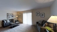This single-floor space centers around a comfortable living room finished with wall-to-wall carpeting that adds warmth underfoot. The room is anchored by a large sliding glass door dressed in vertical blinds, allowing natural light to filter in while offering the option for privacy. The neutral, light-toned walls provide a clean backdrop that complements the space's understated aesthetic.

Climate control is well-considered, with a wall-mounted air conditioning unit positioned on one side of the room alongside baseboard heating along the perimeter — ensuring year-round comfort. A floor lamp provides supplemental ambient lighting, adding a cozy atmosphere to the space.

The living area is furnished with a gray sofa accented with decorative pillows and a sleek glass-top coffee table. Two framed geometric prints displayed on the wall introduce a contemporary design element, giving the room a polished, cohesive look. The overall ambiance is calm and functional, with a neutral color palette throughout that offers a versatile foundation for personalization.