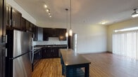 This one-bedroom, one-bathroom unit in Spring, TX is designed with an open, functional layout that maximizes its well-proportioned footprint. The kitchen is a standout feature, outfitted with rich dark cabinetry extending to elevated heights, sleek granite-style countertops, and a full suite of stainless steel appliances including a gas range, microwave, refrigerator, and dishwasher. A center island with pendant lighting above adds both prep space and a natural gathering point that flows seamlessly into the living and dining areas. Wood-style plank flooring runs throughout the main living spaces, lending warmth and continuity, while an arched doorway provides an elegant transition toward the bedroom wing. The living room is generously sized with a ceiling fan, large windows, and access to a balcony, filling the space with natural light and outdoor views. The bedroom features plush carpeting, a bright window with exterior views, and ample square footage for comfortable furnishing. The bathroom includes a soaking tub with shower combo, dark vanity cabinetry, and direct access to a spacious walk-in closet complete with built-in shelving for organized storage. Additional closet space is also available in the living area. The overall ambiance is clean, modern, and move-in ready.
