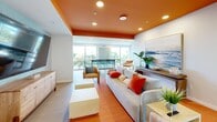 Located at 96 Coco Plum Dr in Marathon, FL, this single-floor space offers a thoughtfully designed combined living and dining area with a distinctly coastal Florida character. The living area is immediately eye-catching, featuring a bold orange accent ceiling that contrasts with crisp white walls and light wood-tone flooring throughout. Comfortable seating, including a spacious grey sofa accented with coral and neutral throw pillows, is arranged around a large flat-screen television mounted against a softly patterned blue-green accent wall. A wooden media console and open shelving add functional storage while maintaining the relaxed, beachy aesthetic. Large windows along the perimeter flood the space with natural light and provide views of the lush surrounding landscape. The dining area features a round white table with aqua wire chairs, contributing a playful, tropical feel. Glass-paneled interior walls and doors in the dining zone create visual openness and connectivity between areas. A small service counter with cabinetry is tucked nearby, adding practical utility. The overall ambiance is bright, welcoming, and distinctly Keys-inspired, blending vibrant color with casual, airy design elements suited to the warm South Florida environment.