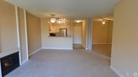 This single-floor space features an open, flowing layout that integrates the living area, kitchen, and an adjoining room. The living area is finished with wall-to-wall carpeting in a neutral tone, creating a clean and understated foundation throughout the space. A ceiling fan with a multi-bulb light fixture illuminates the main living area, providing both functional lighting and air circulation.

The kitchen is positioned along the back wall and is partially open to the living space via a raised pass-through counter with a granite or stone-style surface, ideal for casual dining or food preparation. The kitchen is equipped with a stainless steel refrigerator, microwave, and light-toned cabinetry, offering a practical and organized cooking environment. A doorway adjacent to the kitchen leads to an additional area, likely a utility or entry space.

To the right, a sliding door — potentially a mirrored closet or room divider — opens to a secondary space, also carpeted and fitted with its own ceiling fan. The walls throughout are painted in a warm beige tone, and the textured ceiling contributes a consistent finish across the entire unit.