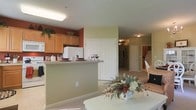 This single-floor residence offers a practical and welcoming layout designed for comfortable everyday living. The kitchen is a standout feature, showcasing warm honey-toned wood cabinetry that extends to the upper walls, complemented by a bold accent wall in deep red that adds a touch of character. A raised breakfast bar serves as a natural divider between the kitchen and the adjacent living and dining areas, creating an open-concept flow while maintaining distinct functional zones. White appliances and light countertops keep the kitchen feeling bright and clean. A large paneled closet door adds storage convenience just off the main living area. The flooring transitions seamlessly from the kitchen area into the carpeted living spaces beyond, contributing to a cohesive and unified interior. The hallway leading further into the residence hints at additional rooms, with ceiling-mounted light fixtures providing consistent, even illumination throughout. The overall ambiance is warm and residential, with a neutral color palette punctuated by rich accent tones, making the space feel inviting and well-balanced. The open sightlines across the main living area enhance the sense of spaciousness throughout the home.