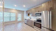 This single-floor residence showcases a clean, contemporary design with a cohesive neutral palette and light hardwood-style flooring that extends throughout the living areas. The kitchen serves as a functional centerpiece, featuring flat-front cabinetry in a warm wood-tone finish paired with crisp white quartz countertops and a classic subway tile backsplash. A full suite of stainless steel appliances—including a French door refrigerator, electric range, over-the-range microwave, and dishwasher—equips the space for everyday use. A matte black faucet at the undermount sink adds a modern accent, while recessed lighting and a glass-paneled exterior door bring in natural light. The open layout connects the kitchen to the main living area, which is bright and versatile, enhanced by a ceiling fan and natural light from a large window with blinds. A dedicated bedroom offers a serene, airy feel with its own window overlooking the exterior. The bathroom is finished with tile flooring, a tub-shower combo with white tile surround, a framed mirror, wood-tone vanity, and coordinating matte black fixtures throughout, tying the design together cohesively.