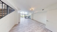 Located at 4400 West Sunrise Boulevard in Plantation, FL, this modern two-story unit offers a well-designed layout spread across two floors. The main level features an expansive open-concept living room and kitchen that immediately impresses with soaring floor-to-ceiling windows, flooding the space with abundant natural light and offering views of lush tropical surroundings, including palm trees and a balcony. Light wood-style flooring runs throughout the living area, complementing the clean white walls and creating a bright, airy atmosphere. The kitchen is outfitted with sleek white cabinetry, dark stone countertops, stainless steel appliances including a refrigerator, electric range, built-in microwave, and a tile backsplash — combining functionality with a contemporary aesthetic. A stylish staircase with dark metal railings leads to the upper level, which provides additional living space, a dedicated closet with built-in wire shelving, and continued light wood flooring. Both bathrooms are finished with large-format marble-style tile flooring, floating white vanities with dark countertops, and glass-enclosed showers, reflecting a clean, modern design sensibility throughout. The overall ambiance is crisp, bright, and minimalist, making this a refined and functional living space.