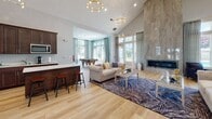 Located at 520 Washington Ave in Dumont, NJ, this single-floor clubhouse-style space is designed with a refined yet inviting aesthetic that balances comfort and functionality. The interior is characterized by dramatic vaulted ceilings that lend an expansive, airy quality to the open-plan layout, with light hardwood flooring running throughout to unify the various zones seamlessly.

The main lounge area is appointed with tufted upholstered seating, layered rugs, and a boldly paneled white accent wall that serves as a focal point, complete with a wall-mounted flat-screen television and stylish side consoles. Gold-toned sputnik and hoop chandeliers add a contemporary elegance overhead. Large windows dressed with patterned drapes invite abundant natural light and offer views of the surrounding greenery.

Adjacent to the lounge, a full-length stone fireplace wall anchors a secondary seating area, providing a dramatic architectural statement. A well-equipped kitchenette with dark cabinetry, stainless steel appliances, a breakfast bar with stools, and light countertops supports gatherings and events. A dedicated game area featuring a billiards table rounds out the space, while glass-paneled doors connect seamlessly to an outdoor area, enhancing the flow between interior and exterior environments.