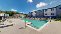 Located at 900 Elm Grove Ave in Gainesville, Georgia, this outdoor amenity area is centered around a generously sized resort-style swimming pool with crystal-clear water and multiple entry points, including steps and stainless steel handrails. The pool deck is expansive, surfaced in smooth concrete, and lined with an extensive collection of lounge chairs and chaise lounges for sunbathing — accommodating a large number of residents comfortably. Shade umbrellas are positioned throughout the deck, offering relief from the Georgia sun.

A standout architectural feature is the covered pavilion adjacent to the pool, constructed with bold brick columns and warm, exposed heavy timber framing beneath a pitched roof. This sheltered space includes outdoor seating and dining furniture, creating an inviting area for relaxation and socializing. The pavilion's craftsman-inspired design adds a refined, lodge-like character to the outdoor setting.

The pool area is enclosed by black iron fencing, providing both security and an unobstructed view of the surrounding landscaping and multi-story residential buildings beyond. Mature trees form a natural backdrop on the perimeter, adding greenery and privacy. The overall ambiance is bright, open, and community-focused, offering a well-appointed outdoor retreat designed for active leisure and casual gatherings under the open Georgia sky.