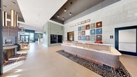 Located in Chicago's West Loop neighborhood, this expansive residential lobby at 1114 West Carroll Avenue presents a thoughtfully designed, single-floor space that makes a striking first impression. The reception area anchors the lobby with a substantial wood-and-stone desk accented by brass detailing, set atop a geometric patterned rug. A curated grid of colorful botanical artwork adorns the concrete accent wall behind the desk, while sleek pendant lighting illuminates the double-height ceiling above. The lounge area is defined by floor-to-ceiling glass curtain walls that flood the interior with natural light and frame views of the surrounding Chicago streetscape. Tall cylindrical bronze pendant lights hang dramatically over a central conference-style table and a well-appointed seating arrangement featuring eclectic, design-forward furniture. A full-height open shelving unit serves as a stylish room divider, displaying books and décor. The mailroom area continues the refined aesthetic with sage green paneling, warm pendant lighting, and curated artwork. Polished concrete floors flow seamlessly throughout, and black-framed glass entry doors complete the contemporary yet inviting character of this well-appointed lobby.