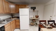 This single-story, 2-bedroom, 1-bathroom home features a practical and well-organized layout designed for efficient everyday living. The kitchen is a standout functional space, outfitted with warm honey-toned wooden cabinetry that provides generous storage throughout. Upper and lower cabinets line the walls, offering ample room for pantry essentials and cookware. A full-size white top-freezer refrigerator anchors the kitchen, complemented by a gas or electric cooktop visible along the counter. The countertops feature a neutral laminate surface that pairs well with the cabinetry. Light-toned flooring runs throughout, contributing to a clean and cohesive aesthetic. A freestanding metal wire shelving unit near the dining area provides additional storage and display space, styled with decorative baskets and accessories. A dining area is situated adjacent to the kitchen, accommodating a table and chairs for casual meals. The overall ambiance of the home is warm and understated, with neutral tones and practical finishes that create a welcoming, livable environment suited to a variety of lifestyles.