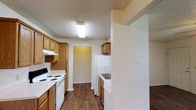 Located at 100 Chapelridge Dr in Richland, Mississippi, this single-story residence offers a practical and well-organized living space. The kitchen is a standout feature, designed with a galley-style layout that maximizes efficiency. Warm-toned wood cabinetry lines both sides of the kitchen, providing ample storage space, while white appliances — including a refrigerator, electric range, and dishwasher — offer a clean, cohesive look. A built-in sink with overhead cabinetry adds to the kitchen's functional design. The textured ceiling and overhead fluorescent lighting ensure the space is well-lit. Dark wood-look flooring runs throughout, adding warmth and durability underfoot. A partial wall divides the kitchen from the adjacent living area, creating a sense of separation while maintaining an open feel between spaces. A doorway at the end of the kitchen leads further into the home, hinting at additional rooms beyond. The overall design reflects a straightforward, functional aesthetic well-suited for comfortable everyday living.