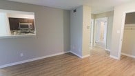 This single-story space presents a clean, well-maintained interior with a neutral color palette that creates a calm and welcoming atmosphere. The main living area features warm wood-look laminate flooring that extends throughout, complementing the soft gray walls and crisp white baseboards for a cohesive, modern aesthetic.

A structural column partially divides the living area, adding architectural character while maintaining an open feel. A pass-through window connects the living space to the kitchen, which is visible in the background and features dark cabinetry, a tiled backsplash, and a countertop that facilitates easy interaction between the two spaces.

A hallway extends toward the rear of the unit, leading to additional rooms visible in the distance. The corridor is well-lit and maintains the same consistent flooring and wall tones. A thermostat is mounted along the hallway wall, and ceiling vents are positioned throughout for efficient climate control.

Overall, the space offers a straightforward, functional layout with updated finishes, making it well-suited for a variety of uses. The consistent use of neutral tones and warm flooring contributes to a bright, inviting environment.