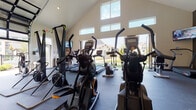 Located at 277 Bend Blvd in Westerville, Ohio, this single-floor fitness center is designed to accommodate a wide range of workout styles and fitness goals. The facility boasts soaring vaulted ceilings that lend an open, airy feel throughout the space, complemented by an abundance of natural light streaming in through large clerestory windows and expansive glass garage-style doors that roll up to connect the interior with the outdoors.

The cardio zone is well-appointed with a lineup of treadmills, elliptical trainers, stair climbers, and rowing machines — many positioned to take advantage of the outdoor views. A dedicated strength training area features a Rogue power rack with barbells and bumper plates, multiple weight benches, and a comprehensive dumbbell rack. Kettlebells and additional functional training tools round out the free weights section.

A turf training lane runs along one side of the facility, ideal for sled pushes, agility drills, and dynamic movements, with Rogue sled attachments and stacked plyo boxes nearby. Rubber flooring throughout provides durability and safety underfoot. Mounted TVs are positioned for viewing from multiple workout stations. The facility also includes a pegboard climbing wall for upper-body conditioning, adding a unique functional training element to this comprehensive community fitness space.