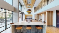 This contemporary amenity lounge is designed with an open, flowing layout that seamlessly connects multiple distinct zones across a single level. At the heart of the space sits a substantial granite-topped island with a textured concrete base, flanked by warm cognac leather bar stools — an inviting centerpiece for casual gatherings or collaborative work. Overhead, a striking arrangement of large dome pendant lights in a bronze finish draws the eye upward toward soaring double-height ceilings accented by a recessed wood-paneled coffer detail.

Adjacent to the island, a large communal dining table with wood chairs occupies a defined area anchored by a natural fiber rug, ideal for group meals or meetings. Expansive floor-to-ceiling glass walls and garage-style doors open directly onto a furnished outdoor terrace, flooding the interior with natural light and creating a seamless indoor-outdoor connection.

The lounge area features generous sectional seating, a sleek wall-mounted fireplace with a stone surround, and a wood slat ceiling detail with pendant globe lighting. A built-in media wall with integrated cabinetry and ambient backlighting adds a refined focal point. Semi-private nook seating defined by vertical wood slat partitions offers quieter retreat options within the larger communal space. Light hardwood flooring unifies the entire layout, complemented throughout by warm wood tones, textured wall finishes, and a cohesive modern palette.