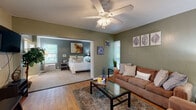 Located at 631 E Call St in Tallahassee, Florida, this single-floor residence offers a well-organized and comfortable layout with a warm, inviting ambiance throughout. The living area is finished with light hardwood flooring and painted in earthy sage green tones, creating a relaxed atmosphere. A ceiling fan overhead, a wall-mounted flat-screen TV, and a glass-topped coffee table anchor the space, complemented by a plush sofa and armchair seating arrangement. The open layout connects the living area seamlessly to the kitchen, which features white cabinetry, a full suite of appliances, a chalkboard accent wall, and a curved breakfast bar with bar stool seating — ideal for casual dining. An arched passthrough enhances the flow between these areas. The bedroom is a calm retreat, dressed in cool blue-gray tones with soft carpeting, a large bed, natural light from exterior-facing windows, and a dedicated desk area suitable for work or study. The bathroom includes a wide vanity with a lighted mirror and ample counter space. Throughout the unit, thoughtful décor, natural light from multiple windows, and cohesive color choices contribute to a comfortable and functional living environment.
