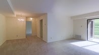 This single-floor residential space presents a clean, neutral interior designed for comfortable everyday living. The main living area is finished with wall-to-wall beige carpet, which continues throughout the unit, creating a cohesive and warm feel underfoot. Crisp white walls provide a bright, open atmosphere, complemented by natural light that streams in from what appears to be a nearby window, casting soft illumination across the floor.

A central partial wall divides the living area, adding subtle architectural definition without fully closing off the space. A hallway extends toward the back of the unit, lined with doors that lead to additional rooms and at least one closet, which features a standard reach-in configuration. An elegant chandelier is visible further down the hallway, adding a decorative touch to the corridor. A wall-mounted heating unit is visible along the baseboard, indicating in-unit climate control. The layout is straightforward and functional, with a logical flow between spaces. The overall ambiance is understated and move-in ready, offering a blank canvas well-suited for personalization.