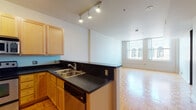 This single-floor residence offers a thoughtfully designed layout that balances function and style. The open-concept kitchen flows seamlessly into the living area, creating a welcoming and versatile space. The kitchen is equipped with light maple cabinetry, dark countertops, stainless steel appliances including an electric range, built-in microwave, refrigerator, and dishwasher, all set atop a classic parquet wood floor. Track lighting overhead provides focused illumination throughout the kitchen and adjoining living space. The living area is bright and airy, benefiting from large windows that frame views of the surrounding urban streetscape, filling the room with natural light. The bedroom continues the city-view theme with oversized windows that look out onto classically styled neighboring architecture. Soft carpeting underfoot adds comfort and warmth to this private retreat. The bathrooms feature clean white tile flooring, light maple vanity cabinetry with dark countertops, and modern fixtures, maintaining a consistent design language throughout the residence. The overall aesthetic is clean and contemporary, with neutral tones, natural materials, and an abundance of natural light defining the character of this urban living space.
