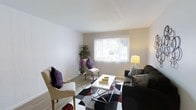 This single-story unit features two bedrooms and two bathrooms within a thoughtfully designed floor plan. The living room serves as the centerpiece of the space, showcasing a clean, contemporary aesthetic with white paneled accent walls that add subtle texture and visual interest. A large window fitted with vertical blinds allows natural light to fill the room, creating a bright and welcoming atmosphere. The living area comfortably accommodates a seating arrangement consisting of cushioned armchairs and a sofa, complemented by a sleek, modern coffee table atop a geometric patterned area rug. Neutral wall tones provide a versatile backdrop that pairs well with bold accent colors introduced through decorative pillows and furnishings. The smooth white ceiling contributes to an open, airy feel throughout. Light-toned flooring runs through the space, enhancing the overall sense of cleanliness and continuity. A decorative plant near the window adds a natural, organic element to the living space. The overall ambiance is calm and contemporary, offering a comfortable everyday living environment suited for a range of lifestyles.