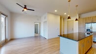 This single-floor residence offers a thoughtfully designed open-concept layout that seamlessly connects the main living areas. Light wood-tone flooring flows continuously throughout the space, contributing to a bright and cohesive aesthetic. The living area is spacious and airy, benefiting from multiple windows dressed with horizontal blinds that allow natural light to filter in, complemented by a ceiling fan for added comfort.

The kitchen is a standout feature, showcasing warm wood cabinetry that extends to the ceiling, a herringbone-pattern tile backsplash with under-cabinet lighting, and dark granite countertops. A large central island with matching granite provides ample prep and dining space, while pendant lighting in gold and black tones adds a contemporary touch. Stainless steel appliances complete the functional and stylish kitchen.

A dining area adjacent to the kitchen is highlighted by an elegant chandelier, adding a refined element to the open floor plan. The bathroom features a walk-in glass-enclosed shower clad in large-format marble-style tile, paired with a wood-toned vanity and dark granite countertop, echoing the design language found throughout the home. The overall ambiance is clean, modern, and move-in ready.
