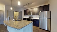 This single-floor residence presents a well-designed open layout that connects the kitchen and living areas cohesively. The kitchen serves as a functional centerpiece, featuring rich dark-espresso cabinetry extending to the upper walls, granite countertops, and a large island with a built-in sink and pendant lighting overhead. A full suite of stainless steel appliances — including a refrigerator, electric range, and over-the-range microwave — completes the space. Subway tile backsplash and track lighting add contemporary detail.

Wood-style plank flooring runs consistently throughout the unit, contributing a warm, unified aesthetic. The living area is accented with crown molding, a ceiling fan, and natural light from multiple windows fitted with blinds. A wall-mounted fireplace niche is visible near the kitchen transition area.

The bedrooms are comfortably sized, with large windows allowing ample natural light and direct hallway access. Two bathrooms mirror the kitchen's design language, each offering dark-framed vanities with granite countertops, framed mirrors, wood-style flooring, and a soaking tub with tiled surround. One bathroom benefits from a bright window, further enhancing the open, airy feel of the space.