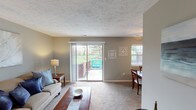 This single-floor residence offers a thoughtfully arranged layout with a comfortable flow between living, dining, and private spaces. The living room is a welcoming focal point, featuring plush carpeting, a textured ceiling, and a large sliding glass door that opens to an outdoor patio and green surroundings, filling the room with natural light. An additional window further brightens the space, which connects seamlessly to the adjacent dining area. The dining room accommodates a full-sized table and chairs beneath a ceiling fan, with its own window providing natural light and a decorative mirror adding visual depth. The galley-style kitchen is efficiently designed with white cabinetry, tile flooring, a full-size refrigerator, dishwasher, and sink, and opens directly into the dining area for easy entertaining. The primary bedroom features a bold navy accent wall, carpet flooring, dual nightstands, and a window with outdoor views, creating a restful retreat. A second bedroom offers softer grey tones, carpet flooring, and a closet. The full bathroom is finished with sage green walls, tile flooring, a shower-tub combination, and a well-lit vanity, giving it a calm, spa-like quality.