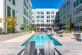 Located at 2230 Calle de Luna in Santa Clara, CA, this generously sized outdoor amenity deck spans an impressive footprint and serves as a central gathering space within a modern multi-story residential community. The space is defined by its resort-style lap pool, which takes center stage and is framed by wide, light-toned tile decking and a series of contemporary lounge chairs ideal for sunbathing. A dedicated spa and shallow wading area complement the main pool, offering varied aquatic experiences.

Shaded retreat zones are provided by elegant wood-and-steel pergola structures, beneath which comfortable seating arrangements invite relaxation and socializing. A fire pit lounge area, furnished with Adirondack-style chairs, provides an inviting setting for cooler evenings. The deck also incorporates thoughtfully designed tiered landscaping with drought-tolerant plantings, raised garden beds, and ornamental trees that add natural texture and color throughout.

The surrounding architecture features a striking multi-story façade with bold yellow and grey paneling and expansive grid windows that overlook the amenity level. Glass railings along the deck's perimeter preserve unobstructed views of the surrounding Santa Clara skyline and distant mountains, while abundant natural light ensures a bright and open atmosphere throughout the day.