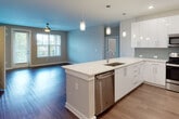 This single-floor residence showcases a thoughtfully designed open-concept layout that seamlessly connects the kitchen and living areas. The kitchen is a standout centerpiece, featuring a large island with a sleek white quartz countertop, an undermount sink, and stainless steel appliances including a French door refrigerator, range, microwave, and dishwasher. Crisp white cabinetry with a high-gloss finish pairs elegantly with a light gray subway tile backsplash, while modern cylindrical pendant lights add a refined touch overhead. Recessed lighting and rich wood-look flooring carry throughout the main living area, which is bathed in natural light from large windows and features a ceiling fan for comfort. The bedrooms offer plush carpeting, ceiling fans, and generous windows with views of lush greenery, creating a calm and restful atmosphere. The primary bathroom is well-appointed with a glass-enclosed walk-in shower, wood-look tile flooring, a white vanity with a quartz countertop, and direct access to a spacious walk-in closet. A secondary bathroom features a tub-shower combination and matching contemporary finishes. The overall aesthetic is clean, modern, and move-in ready, with a cohesive neutral palette accented by soft blue tones.