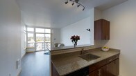 This single-floor space showcases a thoughtfully designed open-concept layout that seamlessly connects the kitchen and living area, creating a bright and cohesive living environment. The kitchen is appointed with rich, warm-toned wood cabinetry, stone granite countertops with subtle variegated patterning, and a stainless steel sink. A breakfast bar extends the counter space, providing a natural transition into the living area and offering an inviting spot for casual dining or entertaining. Track lighting overhead adds a modern touch while ensuring the kitchen is well-illuminated. The living area benefits from generous natural light, courtesy of large floor-to-ceiling windows that frame views of the surrounding neighborhood. A private balcony, accessible through the expansive glazing, extends the living space outdoors and enhances the sense of openness. The flooring throughout is a sleek, dark hardwood that contrasts elegantly with the crisp white walls and ceilings, contributing to the space's clean, contemporary aesthetic. Overall, the unit offers a well-balanced combination of functional design and modern finishes, making it a comfortable and stylish living space.