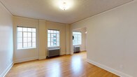 This single-floor space offers a clean, understated interior defined by warm hardwood flooring that runs throughout, reflecting natural light from multiple multi-pane windows along the walls. The windows are generously sized, allowing daylight to fill the open living area and contribute to an airy, welcoming ambiance. Classic cast-iron radiators beneath the windows serve as functional architectural details that nod to traditional building design. The walls are finished in a neutral off-white tone, complementing the light wood floors and providing a versatile backdrop for a variety of interior styles. A textured ceiling adds subtle character overhead, while a central flush-mount light fixture provides additional illumination. An open doorway connects to an adjacent room, also equipped with a radiator and window, suggesting a naturally flowing layout. White baseboards frame the perimeter of the space cleanly. Overall, the interior presents a straightforward, well-maintained environment with timeless architectural elements suited for flexible use.