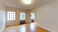 This single-floor space offers a clean, understated interior defined by warm hardwood flooring that runs throughout, reflecting natural light from multiple multi-pane windows along the walls. The windows are generously sized, allowing daylight to fill the open living area and contribute to an airy, welcoming ambiance. Classic cast-iron radiators beneath the windows serve as functional architectural details that nod to traditional building design. The walls are finished in a neutral off-white tone, complementing the light wood floors and providing a versatile backdrop for a variety of interior styles. A textured ceiling adds subtle character overhead, while a central flush-mount light fixture provides additional illumination. An open doorway connects to an adjacent room, also equipped with a radiator and window, suggesting a naturally flowing layout. White baseboards frame the perimeter of the space cleanly. Overall, the interior presents a straightforward, well-maintained environment with timeless architectural elements suited for flexible use.
