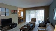 This single-floor residence offers a welcoming and well-lit interior defined by an open layout that seamlessly connects the living and dining areas. The living room serves as the heart of the space, featuring plush carpeted flooring, a dark wood coffee table, and comfortable seating arranged around a central focal point. Large sliding glass doors dressed with vertical blinds flood the room with abundant natural light and provide direct access to an outdoor area, offering pleasant views of surrounding greenery. Decorative wall art adds a pop of color and personality to the neutral-toned walls.

Adjacent to the living room, the dining area is warmly lit by a ceiling fan with integrated lighting and features a dark wood dining table and chairs set on hardwood-style flooring, creating a cozy and inviting atmosphere for meals. A window in the dining area further enhances the sense of openness and allows for additional natural light. The overall ambiance of the space is bright, comfortable, and thoughtfully arranged, blending functional living areas with decorative touches that create a cohesive and inviting home environment.