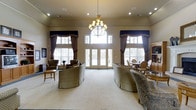 This single-floor space presents a sophisticated and welcoming common area defined by its traditional design sensibility and attention to architectural detail. The room features notably high ceilings that amplify the sense of openness, anchored by a classic multi-arm chandelier that serves as a striking focal point. Recessed lighting complements the natural light that pours in through generously sized windows adorned with navy and taupe valances and full-length drapes, adding a refined, layered look to the space.

A set of elegant French doors at the center of the room opens to the exterior, further enhancing the bright, airy ambiance. The layout is thoughtfully arranged with plush upholstered seating — including curved sofas and accent chairs in neutral tones — positioned to encourage conversation and relaxation. A large area rug grounds the primary seating arrangement, while a smaller sitting nook and a dining area with oval-back chairs can be observed toward the sides of the room. The overall atmosphere is warm and formal, combining classic architectural elements with comfortable furnishings to create an inviting and functional gathering space.