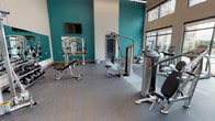 This single-level fitness center located in Corinth, Texas, is designed to support a comprehensive workout experience. The space is characterized by soaring ceilings that create an open, airy atmosphere, complemented by bold teal accent walls that contrast with the neutral white upper walls and ceiling. Large floor-to-ceiling windows line one side of the room, flooding the interior with abundant natural light and providing direct views of an outdoor pool area, enhancing the overall environment.

The flooring throughout consists of durable rubber gym matting in a speckled gray finish, providing both safety and sound absorption. The strength training area is well-appointed with a multi-functional cable machine, a chest press machine, a leg curl/extension machine, an adjustable weight bench, and a dedicated free weight station featuring a tiered dumbbell rack stocked with a full range of weights. A large wall mirror in the free weight area supports proper form during exercise.

The cardio section, situated near the expansive windows, includes treadmills, elliptical machines, and a stationary cycle. Additional accessories such as medicine balls, stability balls, and a wall-mounted television round out the amenity. Linear overhead lighting and well-planned spacing throughout contribute to a functional and comfortable fitness environment.