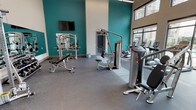 This single-level fitness center located in Corinth, Texas, is designed to support a comprehensive workout experience. The space is characterized by soaring ceilings that create an open, airy atmosphere, complemented by bold teal accent walls that contrast with the neutral white upper walls and ceiling. Large floor-to-ceiling windows line one side of the room, flooding the interior with abundant natural light and providing direct views of an outdoor pool area, enhancing the overall environment.

The flooring throughout consists of durable rubber gym matting in a speckled gray finish, providing both safety and sound absorption. The strength training area is well-appointed with a multi-functional cable machine, a chest press machine, a leg curl/extension machine, an adjustable weight bench, and a dedicated free weight station featuring a tiered dumbbell rack stocked with a full range of weights. A large wall mirror in the free weight area supports proper form during exercise.

The cardio section, situated near the expansive windows, includes treadmills, elliptical machines, and a stationary cycle. Additional accessories such as medicine balls, stability balls, and a wall-mounted television round out the amenity. Linear overhead lighting and well-planned spacing throughout contribute to a functional and comfortable fitness environment.