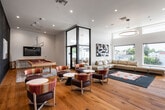 This contemporary single-floor amenity space is designed with a open-concept layout that seamlessly connects multiple functional zones, all unified by warm, wide-plank hardwood flooring throughout. The main lounge area is anchored by a generous sectional sofa, a striking large-scale art piece, and soaring double-height windows that flood the space with natural light and offer expansive views of the surrounding neighborhood. A sleek, modern fireplace with a fluted tile surround and wall-mounted television serves as a focal point in the seating area. The kitchen features deep navy cabinetry extending to the ceiling, a white waterfall-edge island, marble-look backsplash, pendant lighting, and stainless steel appliances — all set against a dramatic dark ceiling with natural light streaming in from adjacent windows. A dedicated games room accommodates a full-size billiards table, wall-mounted television, and bar-height seating, opening directly to an outdoor area through floor-to-ceiling glass doors. Additional lounge seating zones feature richly upholstered chairs in burgundy and multicolor accents arranged around contemporary side tables. A textured dark accent wall provides a bold visual contrast throughout the social gathering areas, and a staircase with a vibrant orange accent wall connects to adjacent levels.