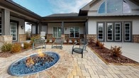 This outdoor amenity space is designed to offer a resort-like experience within a residential community. The exterior of the building showcases a blend of white horizontal siding and stacked stone veneer accents, complemented by a dark metal roof and covered porch areas supported by painted columns. Wall-mounted lantern-style light fixtures add a classic touch to the façade, while large windows allow natural light to flow inside.

The centerpiece of the patio area is a circular fire pit constructed with stone masonry and filled with blue fire glass, creating a striking visual focal point. Surrounding the fire pit are metal wicker-style chairs arranged for social gatherings. The expansive patio itself is paved with multi-toned interlocking stone pavers that extend broadly across the space, connecting various seating zones.

Manicured landscaping featuring ornamental shrubs with warm seasonal foliage lines the building's foundation, adding color and definition. Beyond the patio, a resort-style swimming pool is visible, outfitted with lounge seating, shade umbrellas, and decorative lampposts. The broader community backdrop includes multi-story residential buildings, creating a cohesive, well-planned neighborhood atmosphere.