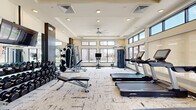 Located in Glendale, Arizona, this modern fitness center offers a thoughtfully designed workout environment packed with a comprehensive range of equipment. The space features a tray ceiling with recessed lighting and a ceiling fan, contributing to a bright and comfortable atmosphere. Large windows with clerestory panels line the walls, flooding the interior with natural light and providing views of the surrounding outdoor amenity areas, including palm trees and community spaces.

The flooring throughout consists of patterned, textured carpet tiles in neutral tones, offering both durability and a contemporary aesthetic. Full-length mirrors are strategically placed along one wall, enhancing the sense of openness and allowing users to monitor their form during workouts.

The cardio zone is well-appointed with multiple Life Fitness treadmills, stair climbers, and a stationary bike with a digital display. The strength training area includes a rack of dumbbells in varying weights, a weight bench, a cable machine, and a multi-function strength station, catering to a full range of resistance training needs. A flat-screen TV is mounted on a central column, providing entertainment during workouts. Dark accent columns and paneling add a refined, modern contrast to the light-toned walls, giving the facility a polished, professional appearance.