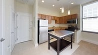 This single-floor residence offers a thoughtfully designed layout combining modern finishes with functional living spaces. The kitchen serves as a central highlight, featuring warm honey-toned cabinetry, granite countertops, and a full suite of stainless steel appliances including a refrigerator, electric range, and microwave. Recessed and pendant lighting illuminate the space, while light tile flooring adds a clean, polished look. A freestanding prep table with a stainless steel top provides additional workspace and storage. The kitchen opens toward the living area, which transitions to plush carpeted flooring, creating a comfortable and inviting atmosphere. Large windows allow ample natural light to fill the space while offering views of the surrounding urban landscape. The bedroom is generously proportioned with soft carpet underfoot, neutral walls, and a set of double doors leading to a closet, offering practical storage. The full bathroom is finished with warm wood vanity cabinetry topped with granite, a single undermount sink, a wall-mounted mirror with overhead lighting, and a full tub-shower combination accented by dark tile detailing. Light tile flooring ties the bathroom together cleanly. Overall, the space presents a cohesive, modern aesthetic suited for comfortable everyday living.