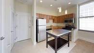This single-floor residence offers a thoughtfully designed layout combining modern finishes with functional living spaces. The kitchen serves as a central highlight, featuring warm honey-toned cabinetry, granite countertops, and a full suite of stainless steel appliances including a refrigerator, electric range, and microwave. Recessed and pendant lighting illuminate the space, while light tile flooring adds a clean, polished look. A freestanding prep table with a stainless steel top provides additional workspace and storage. The kitchen opens toward the living area, which transitions to plush carpeted flooring, creating a comfortable and inviting atmosphere. Large windows allow ample natural light to fill the space while offering views of the surrounding urban landscape. The bedroom is generously proportioned with soft carpet underfoot, neutral walls, and a set of double doors leading to a closet, offering practical storage. The full bathroom is finished with warm wood vanity cabinetry topped with granite, a single undermount sink, a wall-mounted mirror with overhead lighting, and a full tub-shower combination accented by dark tile detailing. Light tile flooring ties the bathroom together cleanly. Overall, the space presents a cohesive, modern aesthetic suited for comfortable everyday living.